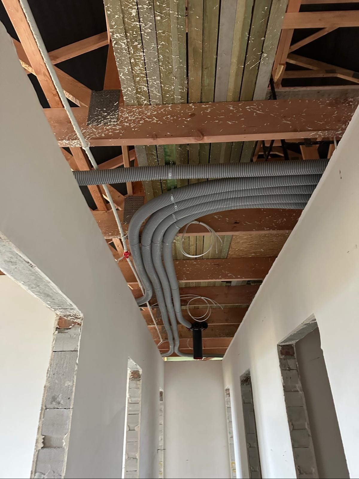 Szare, elastyczne rury wentylacyjne pod drewnianym stropem w surowym wnętrzu. Widoczne fragmenty ścian z cegieł i pustaków. Instalacja w trakcie realizacji.
