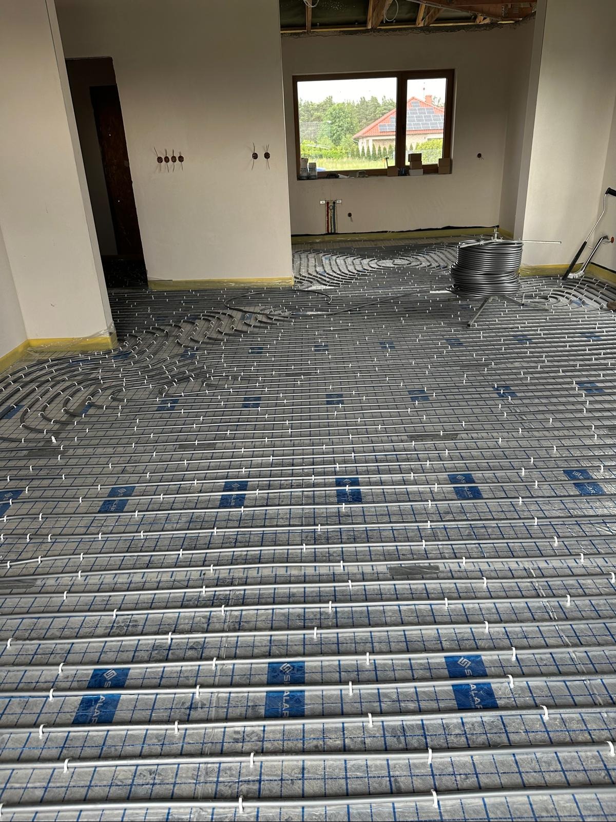 Ułożona instalacja ogrzewania podłogowego w surowym wnętrzu domu, widoczne rury na folii z rastrem, przygotowane do zalania posadzką. W tle okno i fragment instalacji.