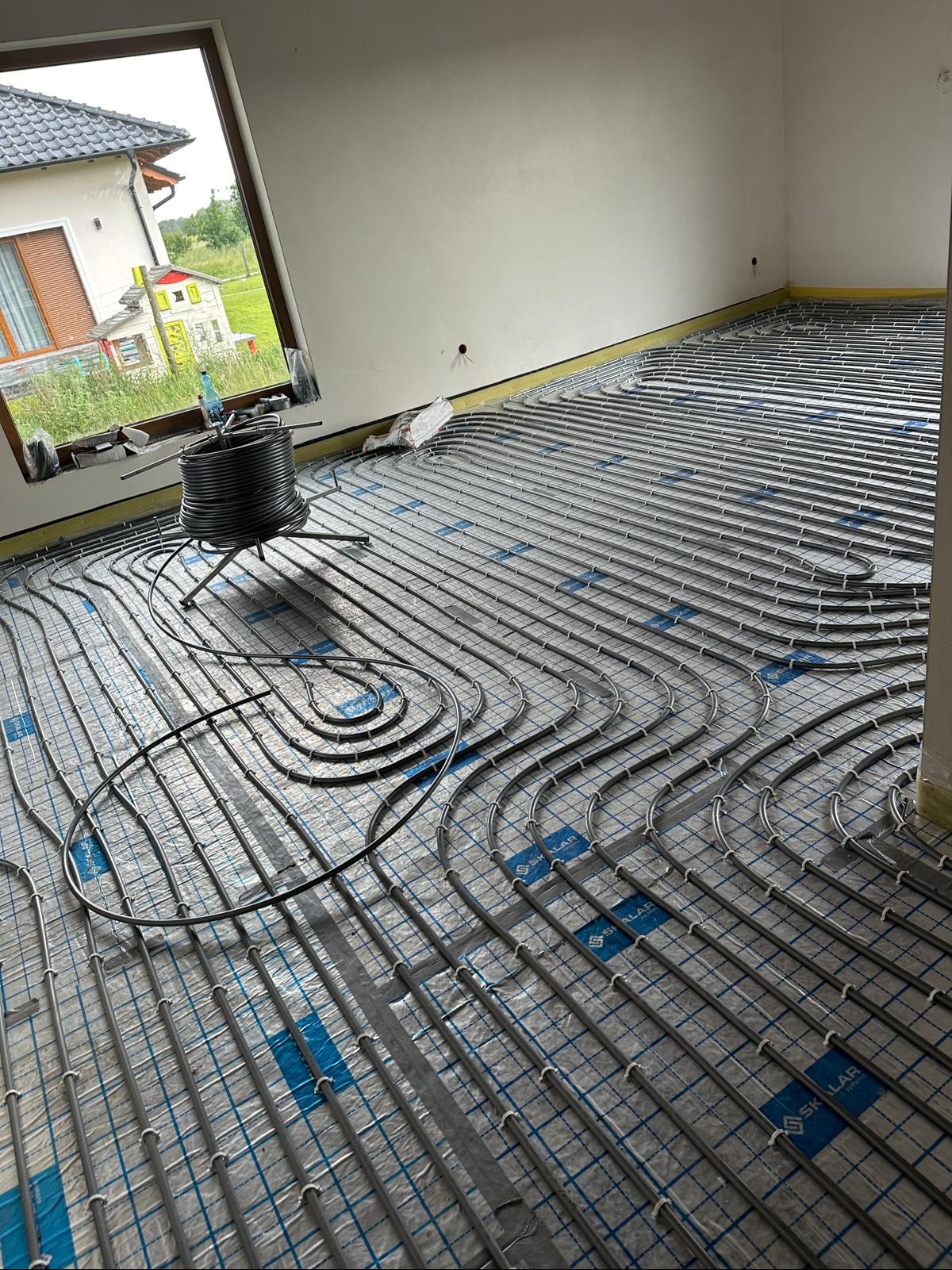 Ułożona instalacja ogrzewania podłogowego na folii z nadrukiem, widoczna cewka z rurą i okno z widokiem na dom. Etap przed wylewką.
