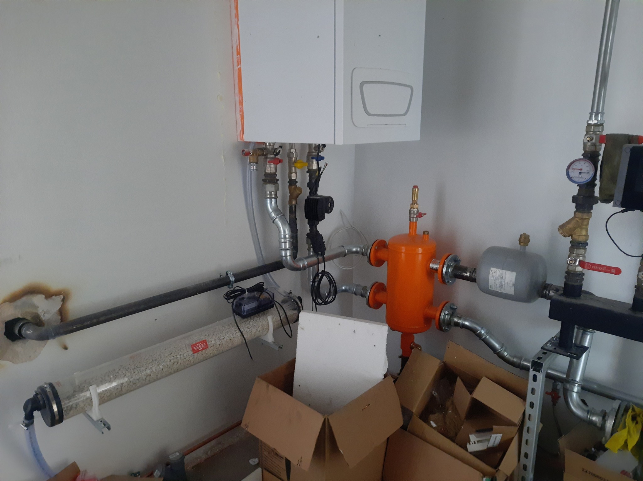 Biały piec CO wiszący nad pomarańczowym filtrem i szarym zbiornikiem wyrównawczym. Widoczne rury, zawory i manometr. W tle biała ściana i kartony.
