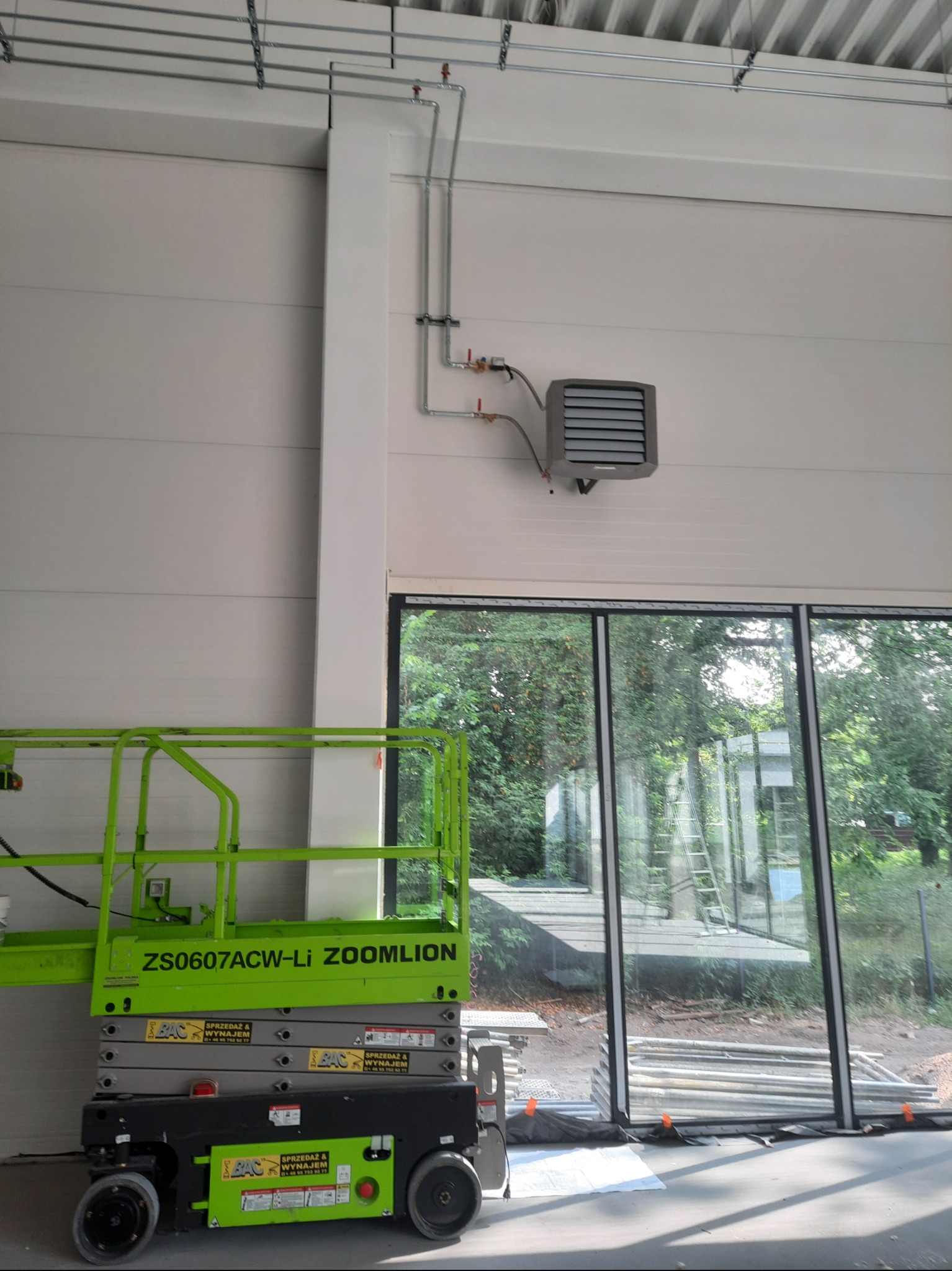 Szara nagrzewnica przemysłowa zamontowana na białej ścianie hali, widoczna z podnośnika nożycowego. Rury instalacji grzewczej prowadzone wzdłuż ściany i sufitu.