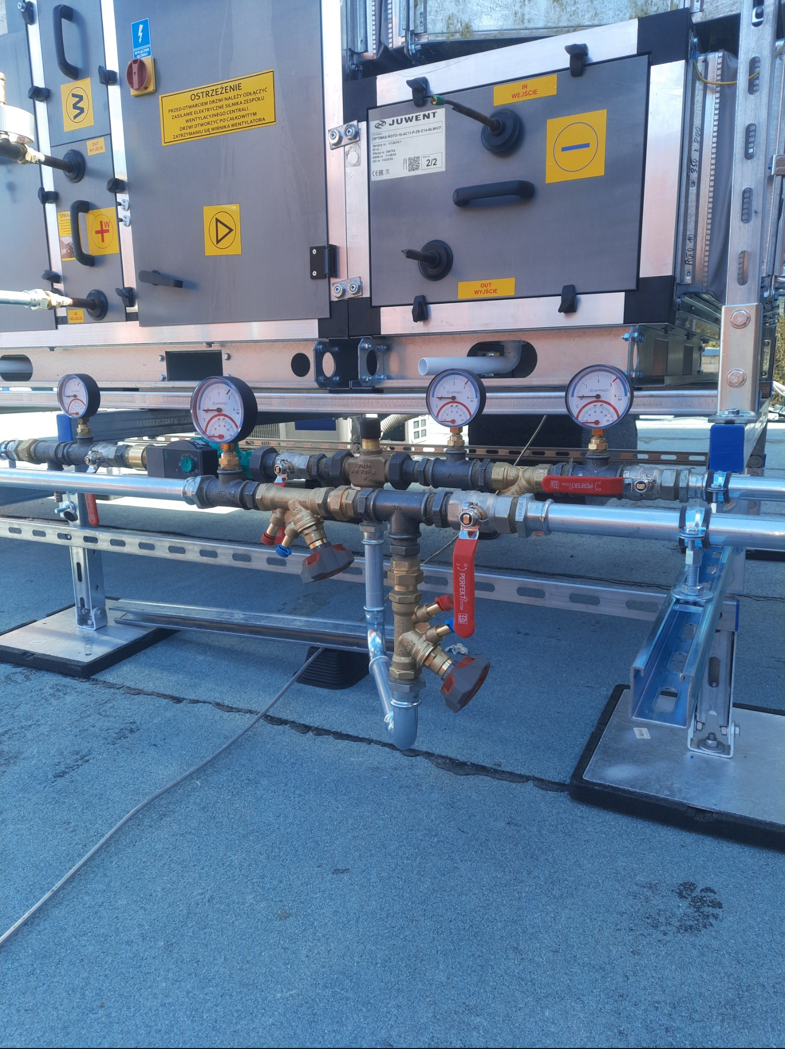 Instalacja przemysłowa z rurami, zaworami i manometrami na metalowej konstrukcji wsporczej. Widoczne oznaczenia ostrzegawcze i tabliczki informacyjne na urządzeniu.