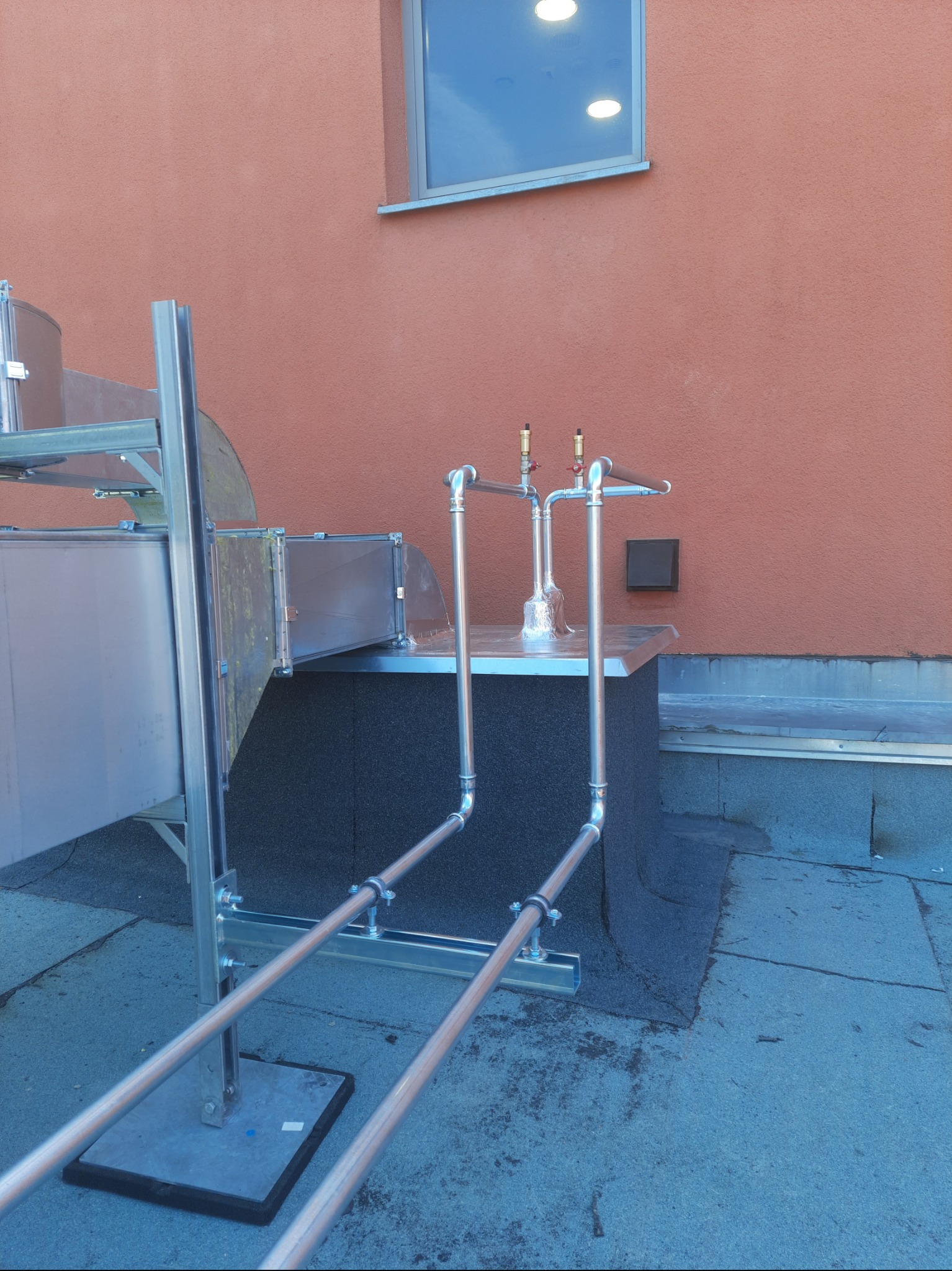 Instalacja wentylacyjna na dachu budynku z pomarańczową ścianą. Widoczne metalowe rury, zawory i elementy mocujące. Ujęcie z dołu, podkreślające złożoność konstrukcji.