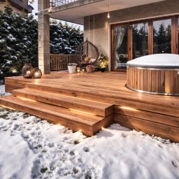 DK spawanie & Budownictwo - Zimowy taras z drewna z jacuzzi i oświetleniem LED w stopniach. W tle dom z balkonem i iglaki pod śniegiem. Dekoracyjne lampiony i fotel wiszący.