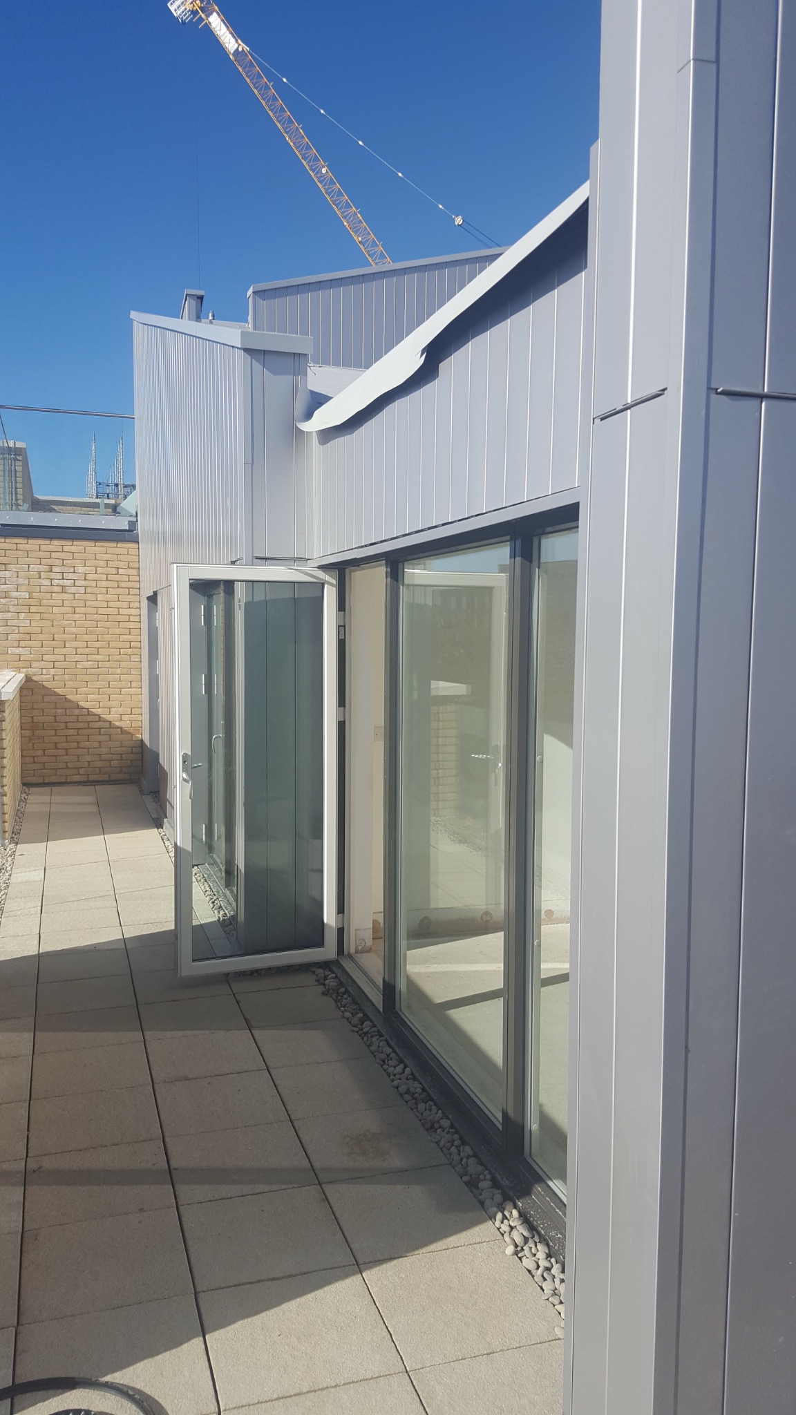 Nowoczesna elewacja budynku z metalowymi panelami i dużymi przesuwnymi oknami, widok z tarasu wyłożonego płytkami. W tle widoczny żuraw budowlany.