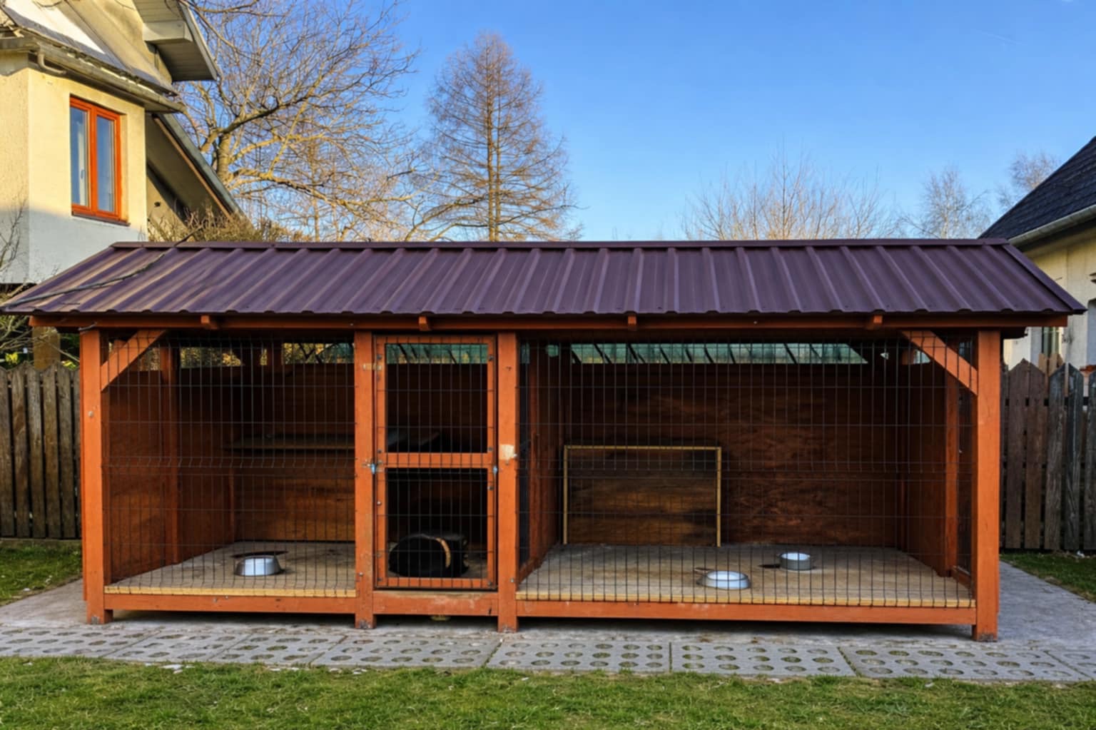 Drewniana konstrukcja z metalowym dachem, prawdopodobnie kojec dla psa, z miskami w środku, na tle domu i drzew.