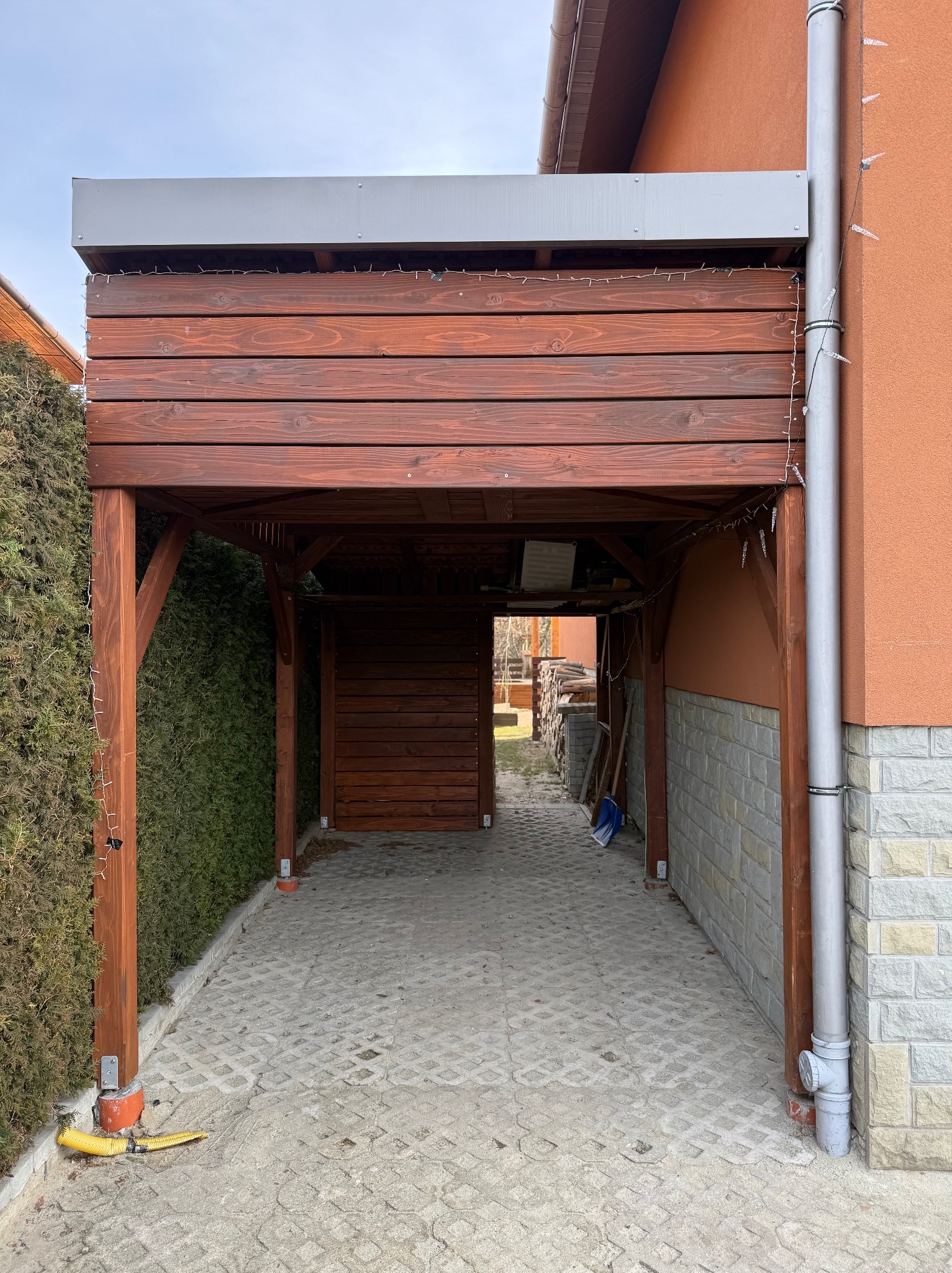 Drewniana pergola z oświetleniem, przylegająca do budynku z elewacją z kamienia. Widoczny fragment ogrodu i kostka brukowa. Konstrukcja z ciemnego drewna.