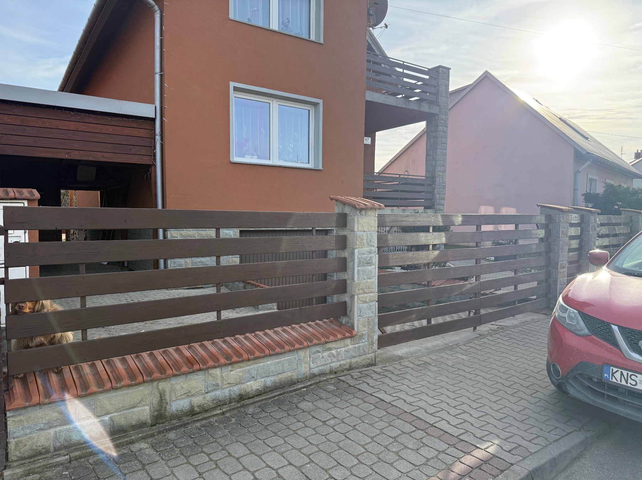 Drewniane ogrodzenie poziome z kamiennymi słupkami i elementami z cegły klinkierowej, przylegające do domu z brązową elewacją. Pies wygląda zza ogrodzenia.