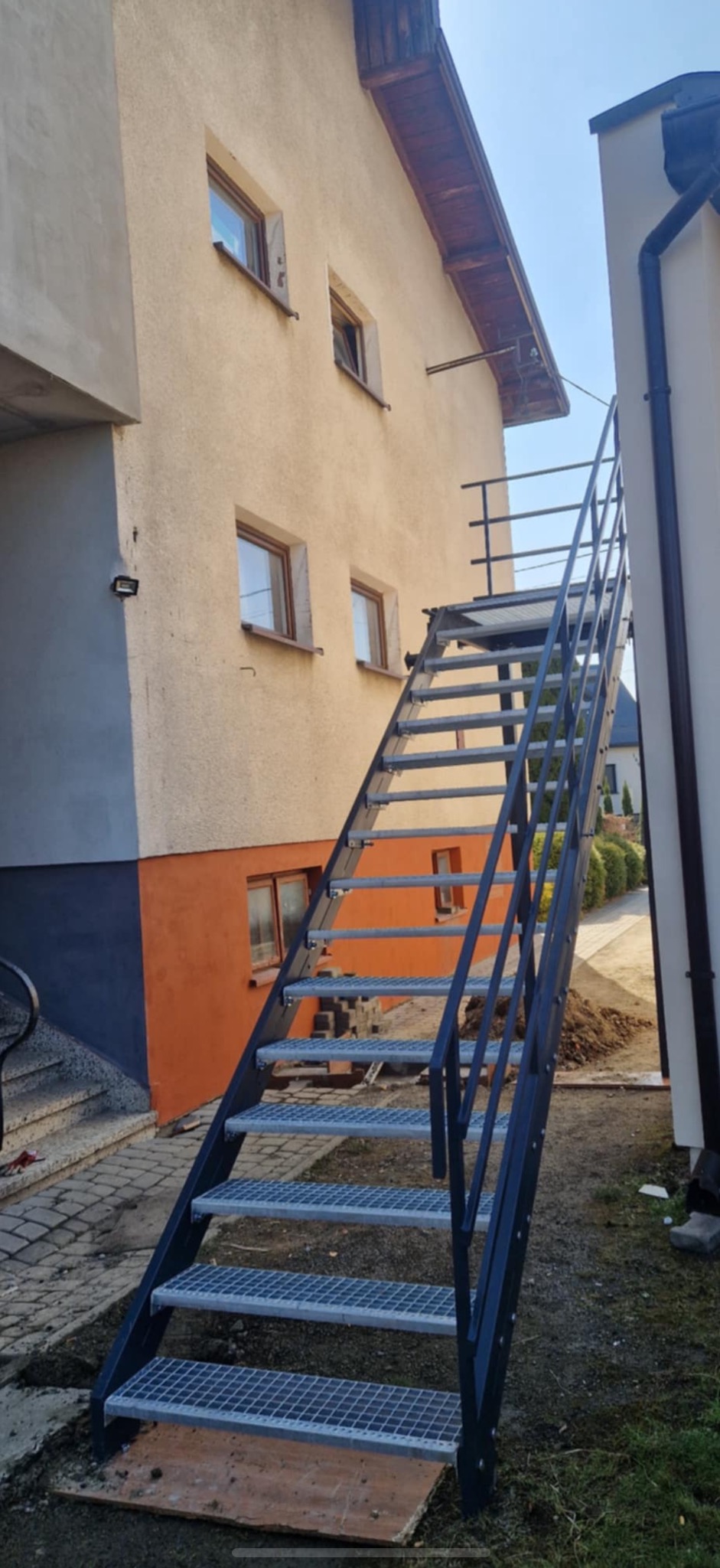 Zewnętrzne metalowe schody z kratowymi stopniami, prowadzące do budynku z oknami. Konstrukcja pomalowana na ciemny kolor, widoczne mocowania do ściany.