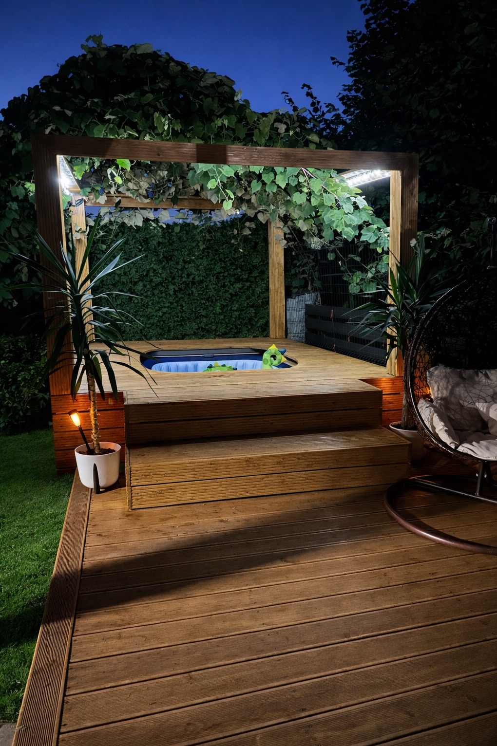 Oświetlona drewniana pergola nad jacuzzi na tarasie z desek, otoczona zielenią w nocy. Rośliny pną się po konstrukcji, tworząc przytulny klimat.