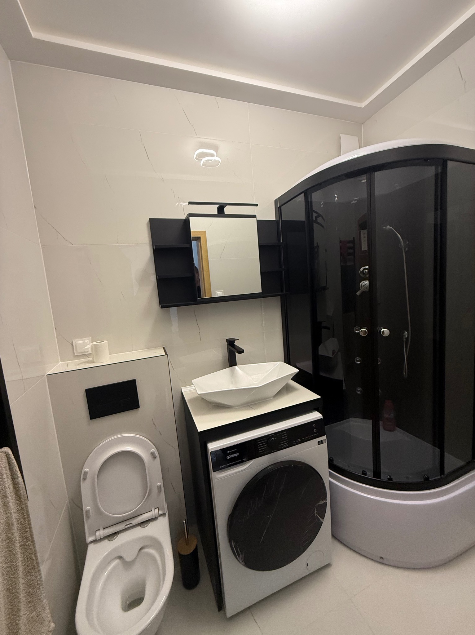 Nowoczesna łazienka z kabiną prysznicową, umywalką na szafce z pralką i podwieszaną toaletą. Białe płytki imitujące marmur, czarne akcenty. Oświetlenie sufitowe.