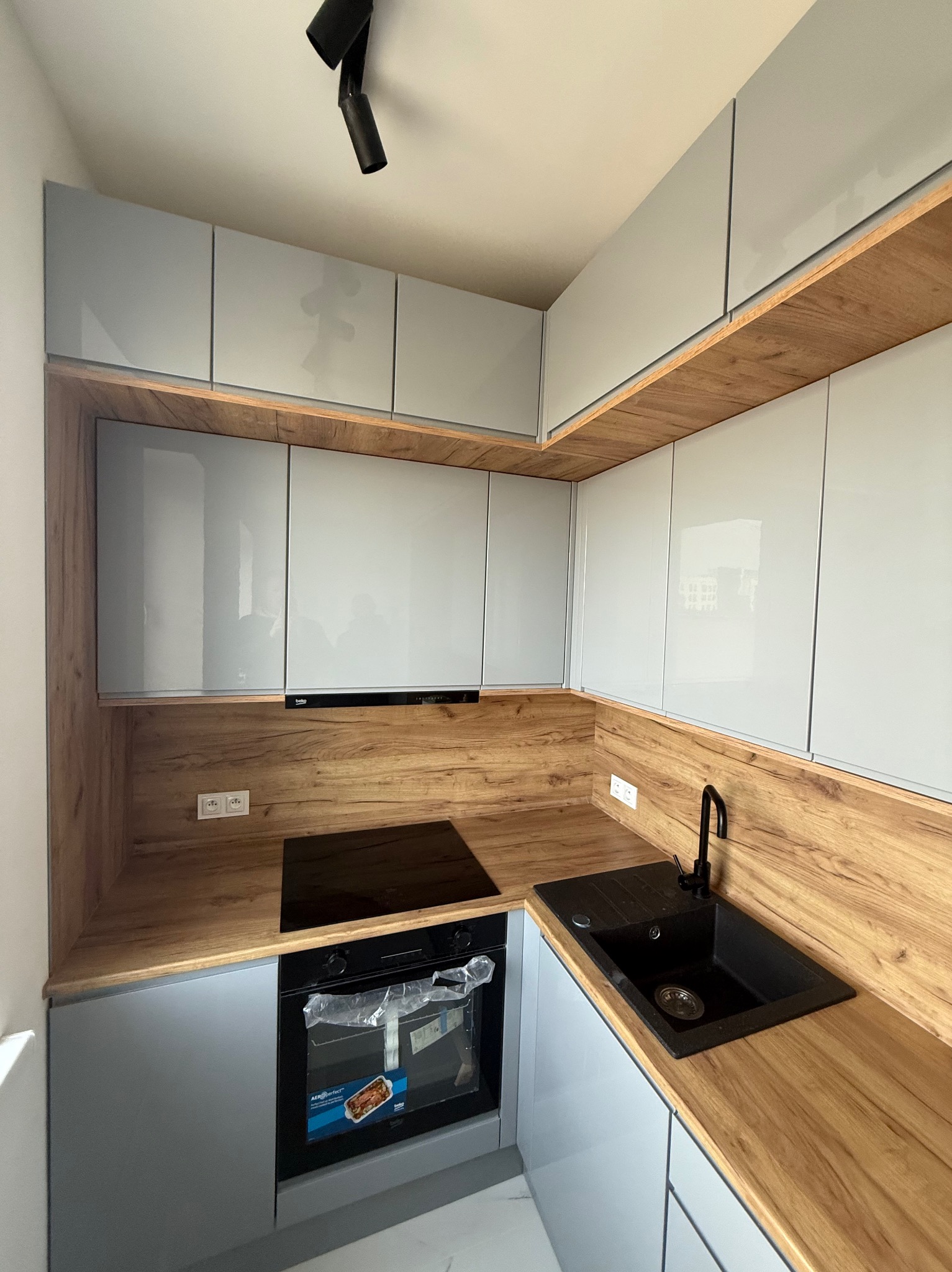 Nowoczesna kuchnia z szarymi szafkami i drewnianym blatem. Czarny zlew i płyta indukcyjna. Oświetlenie punktowe. Minimalistyczny design.