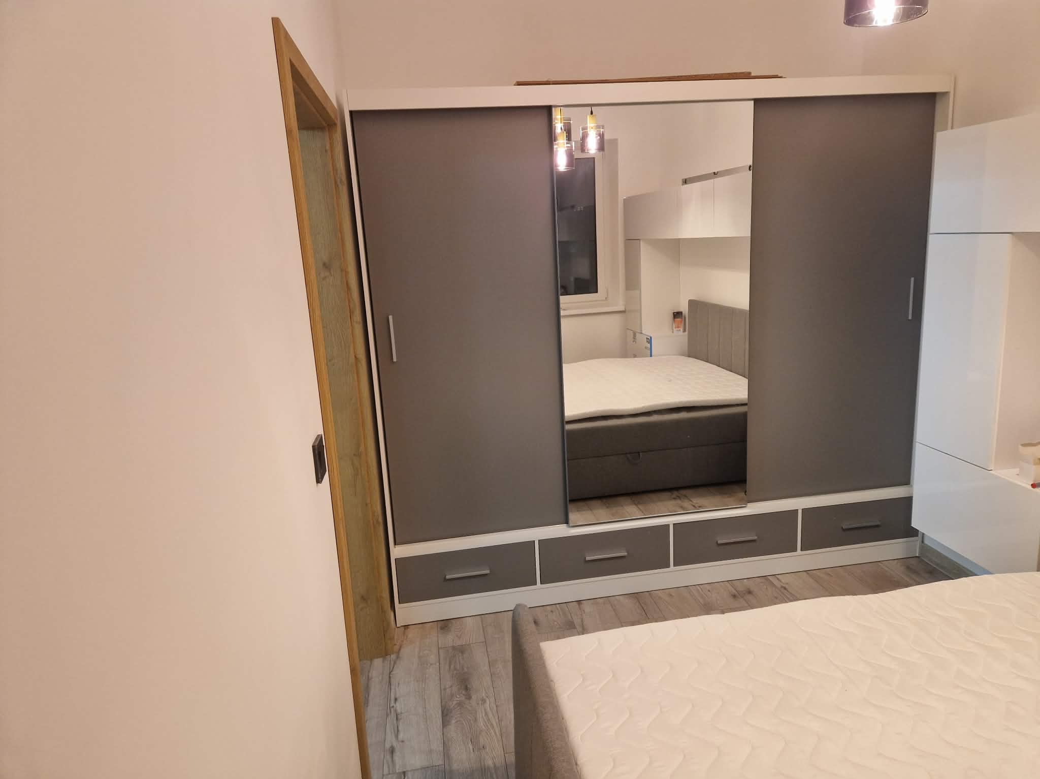 Szafa wnękowa z przesuwnymi, szarymi drzwiami i lustrem, w którym odbija się łóżko. U dołu szafy widoczne szuflady. Całość w jasnym wnętrzu sypialni.