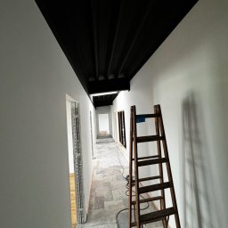 Good-Wall s.c. - Korytarz w trakcie malowania: białe ściany kontrastują z czarnym sufitem. Drewniana drabina oparta o ścianę. Widoczne przygotowania do dalszych prac wykończeniowych.