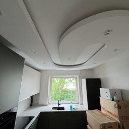 Good-Wall s.c. - Nowoczesna kuchnia z ciemnozielonymi szafkami, białym blatem i oryginalnym sufitem podwieszanym z oświetleniem punktowym. Widok na okno i kartony w tle.