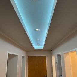 Good-Wall s.c. - Sufit podwieszany z oświetleniem LED w korytarzu, widok z dołu. Centralnie umieszczone drzwi wejściowe, po bokach widoczne wejścia do pomieszczeń. Ściany w jasnym odcieniu.