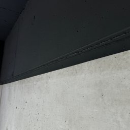Good-Wall s.c. - Fragment ściany: surowy beton w dolnej części i pomalowana na czarno górna część, oddzielone wyraźną linią. Minimalistyczne ujęcie, kontrast faktur i kolorów.