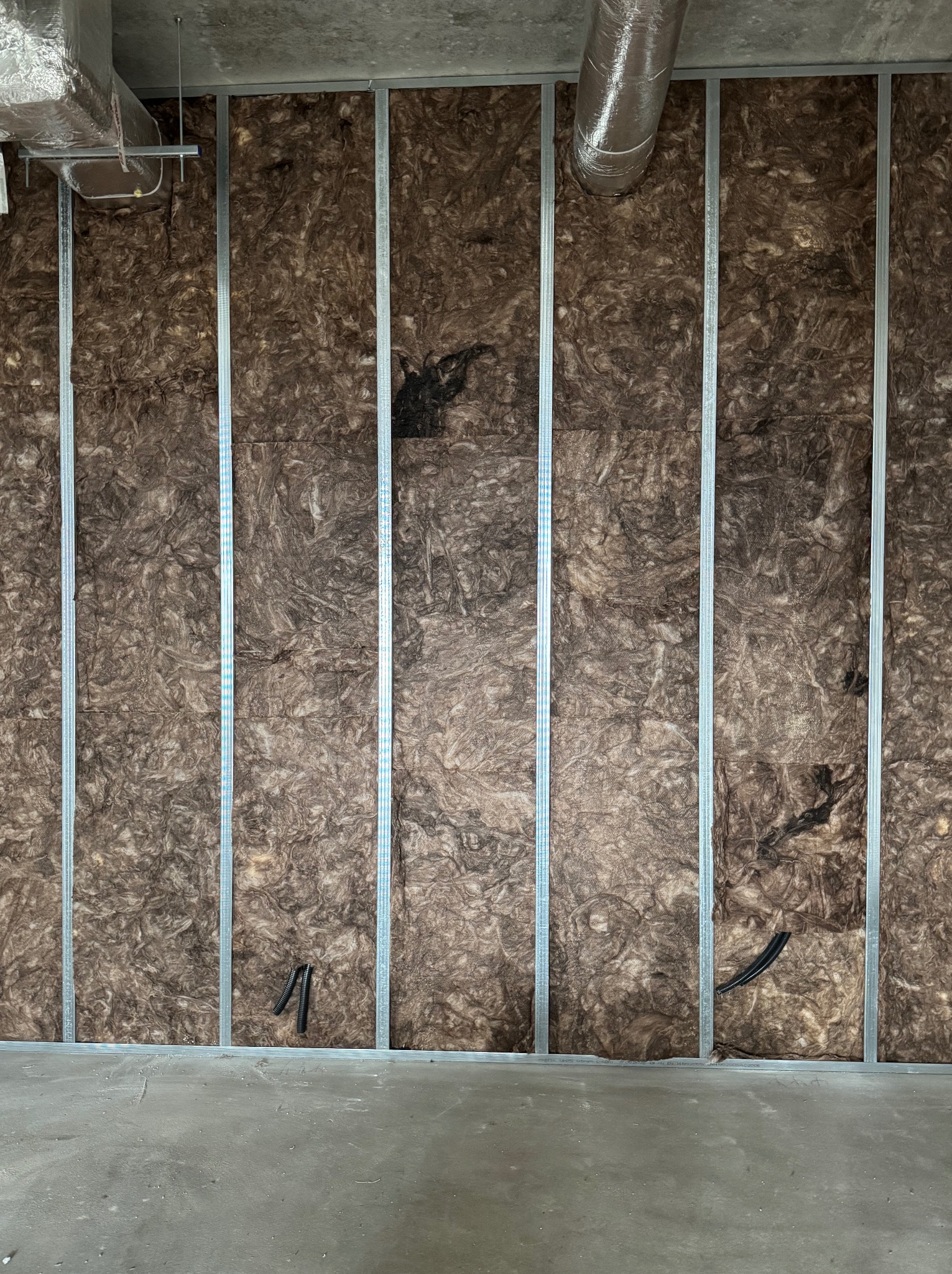 Ściana z izolacją z wełny mineralnej, zabezpieczona metalowym stelażem. Widoczne przewody i elementy wentylacji pod sufitem. Surowy beton na podłodze.