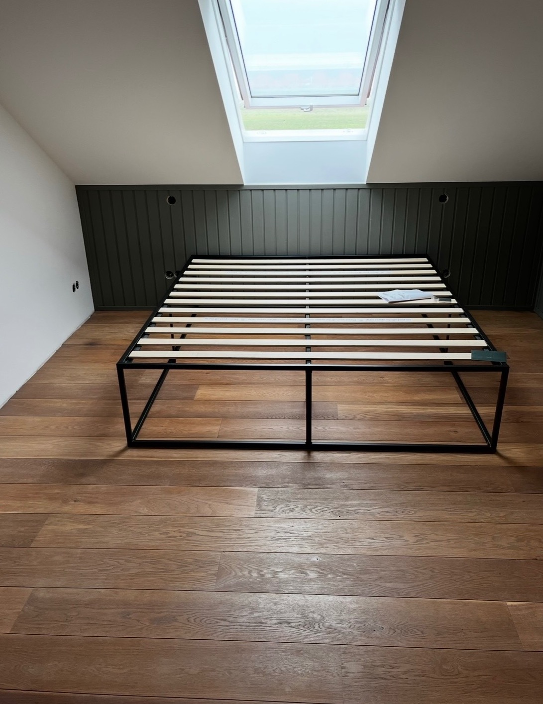 Rama łóżka z drewnianym stelażem na poddaszu z oknem dachowym i drewnianą podłogą. Ciemnozielona boazeria na ścianie.