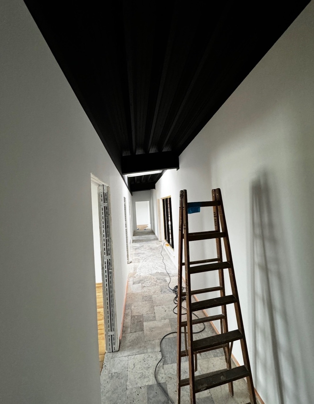 Korytarz w trakcie malowania: białe ściany kontrastują z czarnym sufitem. Drewniana drabina oparta o ścianę. Widoczne przygotowania do dalszych prac wykończeniowych.
