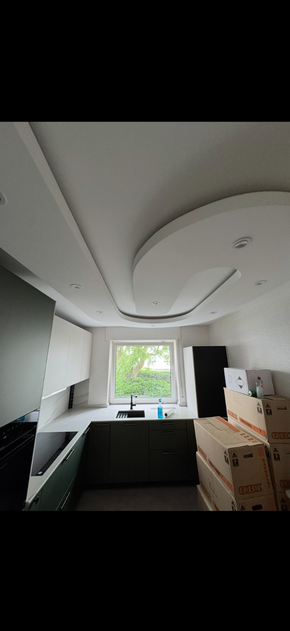 Nowoczesna kuchnia z ciemnozielonymi szafkami, białym blatem i oryginalnym sufitem podwieszanym z oświetleniem punktowym. Widok na okno i kartony w tle.