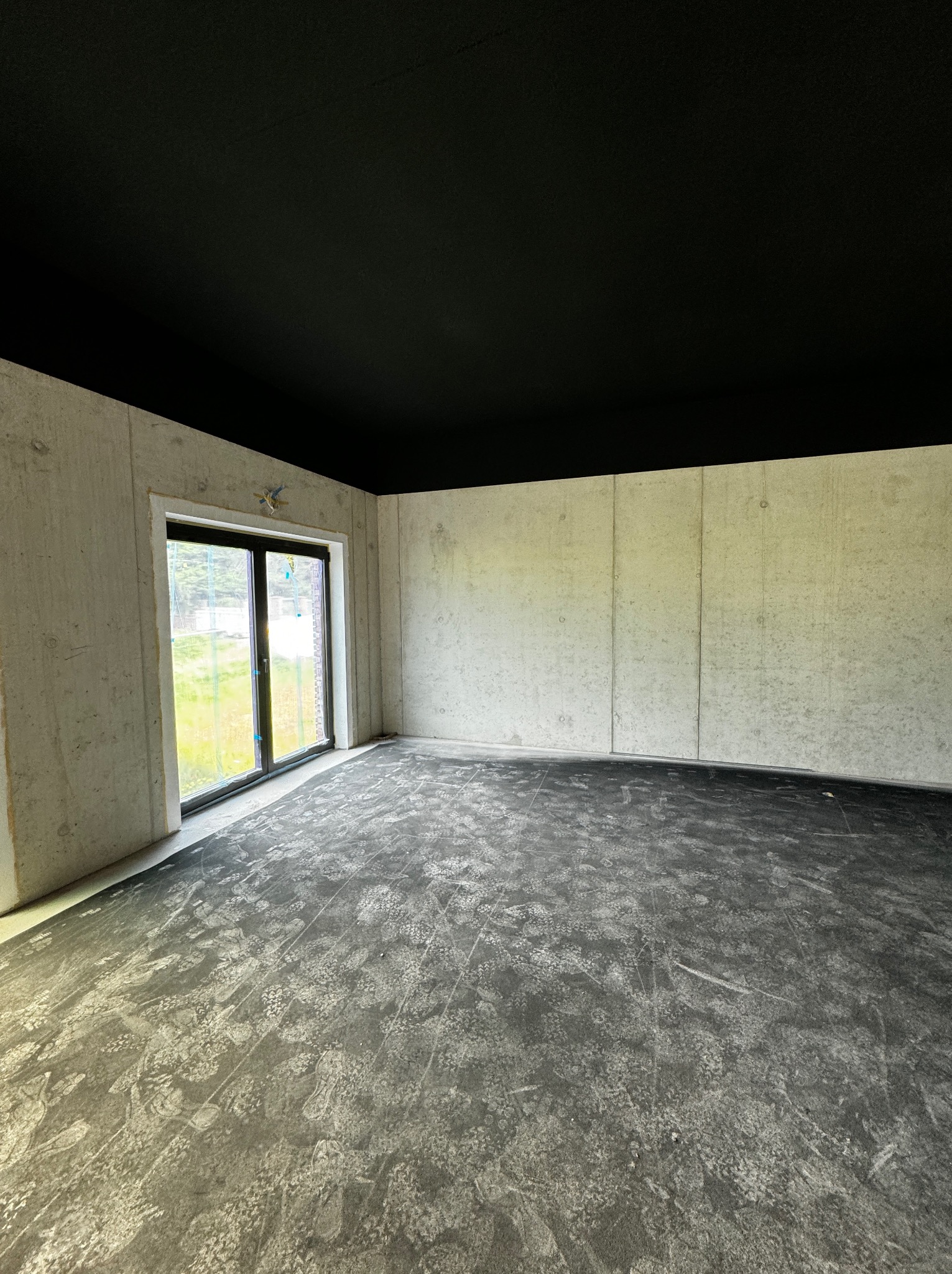 Surowe wnętrze: betonowe ściany, czarny sufit i podłoga z widocznymi śladami. Okno z widokiem na zieleń. Przygotowanie do dalszych prac wykończeniowych.