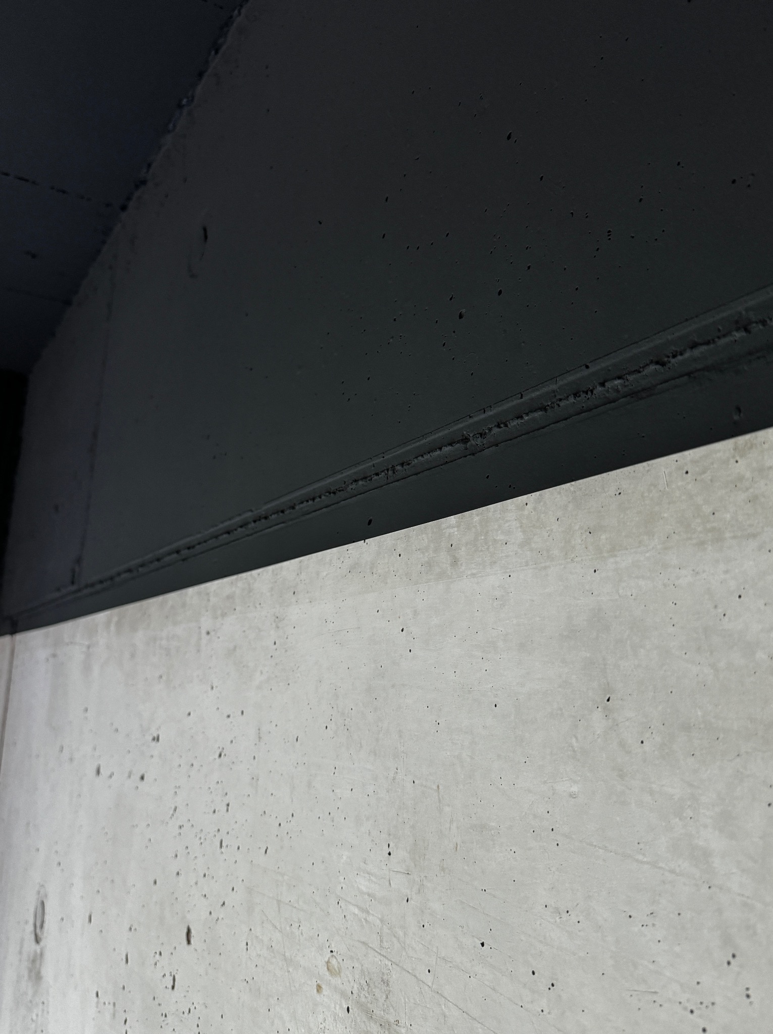 Fragment ściany: surowy beton w dolnej części i pomalowana na czarno górna część, oddzielone wyraźną linią. Minimalistyczne ujęcie, kontrast faktur i kolorów.