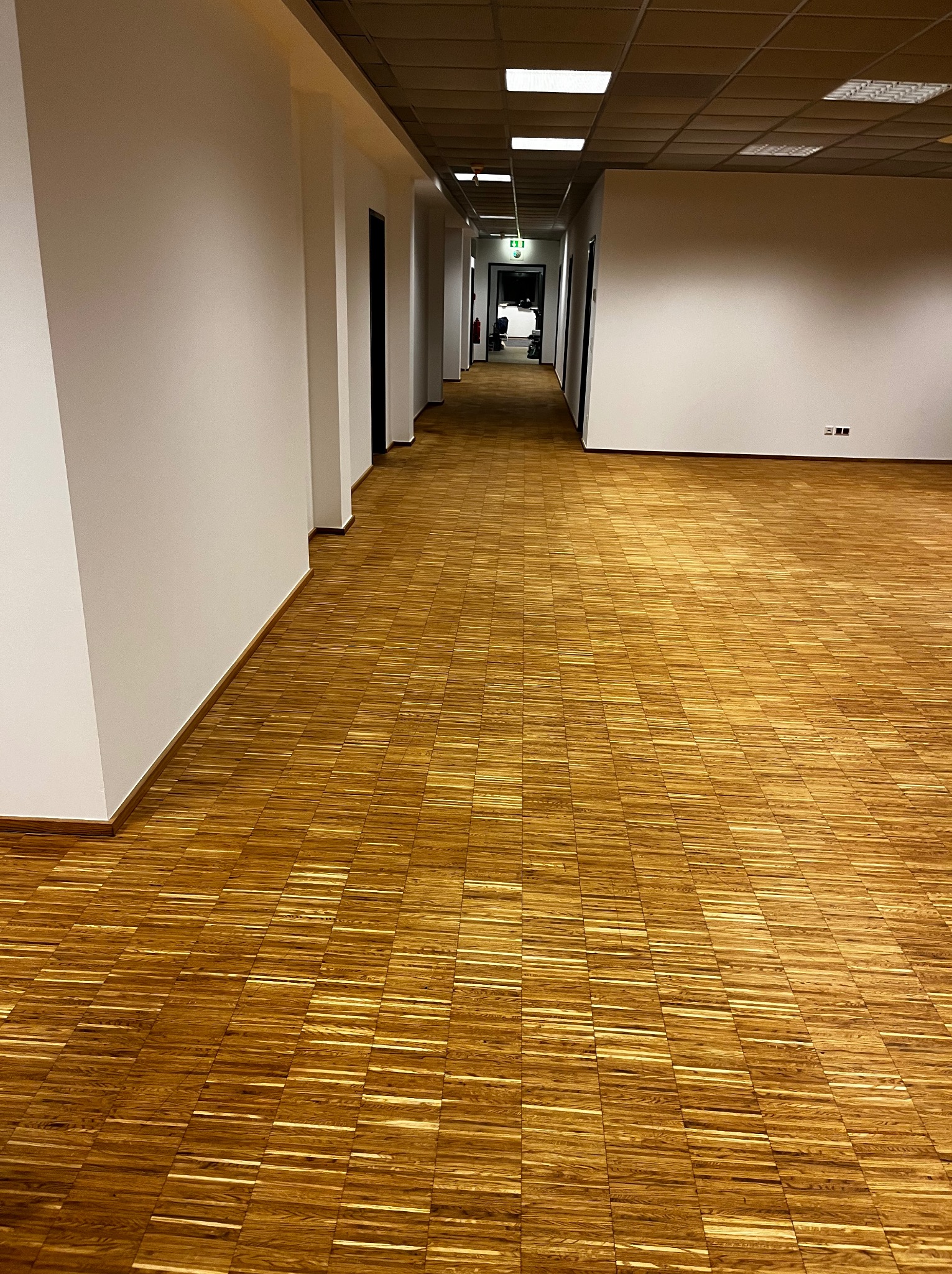 Długi korytarz biurowy z parkietem w jodełkę, świeżo pomalowanymi białymi ścianami i czarnymi futrynami drzwi. Widok w głąb korytarza, oświetlenie sufitowe.