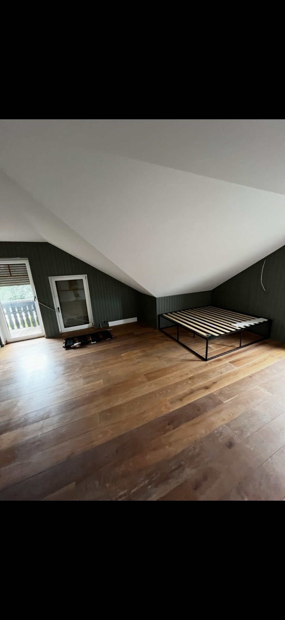 Poddasze po remoncie: świeżo ułożony parkiet z wyraźnym rysunkiem drewna, ciemne panele ścienne i białe skosy sufitu. Rama łóżka bez materaca w rogu pokoju.
