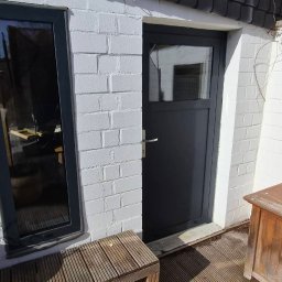 BMHS - Szara stolarka okienna i drzwiowa w białej, ceglanej ścianie zewnętrznej. Widoczny fragment drewnianej ławki i skrzyni. Ujęcie z zewnątrz budynku.