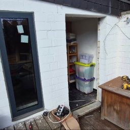 BMHS - Biała ściana z cegły z oknem i metalową kratą. W tle widoczne narzędzia i materiały budowlane, sugerujące prace remontowe w toku.