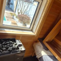 BMHS - Drewniana sauna z piecem na kamienie i oknem dachowym, przez które widać ogród. Wnętrze wykończone jasnym drewnem, na podłodze ciemne płytki. Relaks w domowym spa.