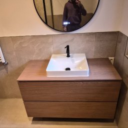 BMHS - Nowoczesna łazienka z drewnianą szafką pod umywalką, białą umywalką i czarną baterią. W tle okrągłe lustro z odbiciem osoby i grzejnik drabinkowy.