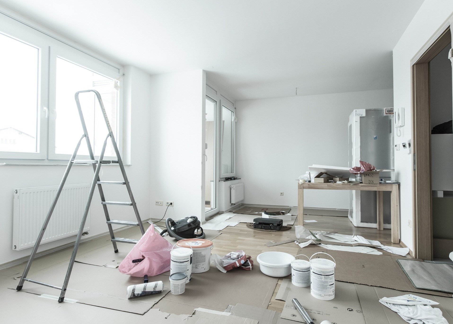 Jasne wnętrze w trakcie malowania: drabina oparta o ścianę, farby, wałek, zabezpieczona podłoga kartonem. Widoczne okno i drzwi balkonowe.