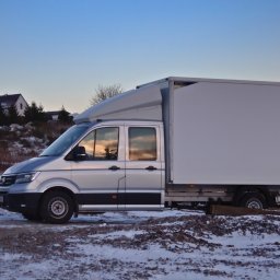 Transport Busem z Windą TBW - Srebrny bus dostawczy z białą zabudową kontenerową na tle zimowego krajobrazu i zachodzącego słońca odbijającego się w szybach. Widok z boku, lekko z dołu.