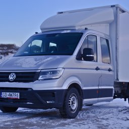 Transport Busem z Windą TBW - Srebrny bus dostawczy VW Crafter z białą zabudową kontenerową na tle zimowego krajobrazu w Skierniewicach. Widoczna tablica rejestracyjna i ośnieżone podłoże.