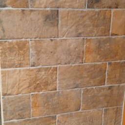 VikRemBud - Ukończona ściana z brązowych, prostokątnych płytek imitujących cegłę, z jasną fugą. Widoczny fragment drewnianej ramy drzwi po prawej stronie.