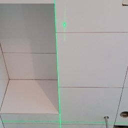 VikRemBud - Białe płytki ścienne w łazience, układane z użyciem lasera krzyżowego. Widoczne akcesoria glazurnicze na podłodze. Ujęcie z dołu, podkreślające precyzję.