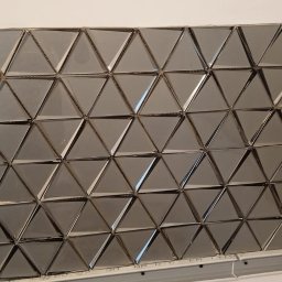 VikRemBud - Ściana wyłożona błyszczącymi, trójkątnymi płytkami w odcieniach szarości, tworzącymi geometryczny wzór. Widoczny fragment ściany bez płytek i puszka elektryczna w Brwinowie.
