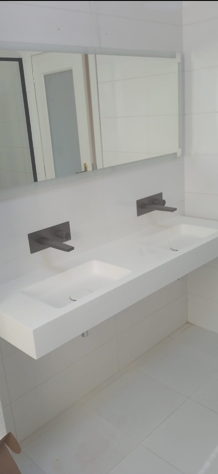 Nowoczesna łazienka z podwójną, białą umywalką nablatową i minimalistyczną, czarną armaturą. Lustro w tle odbija drzwi. Białe płytki na ścianach i podłodze.