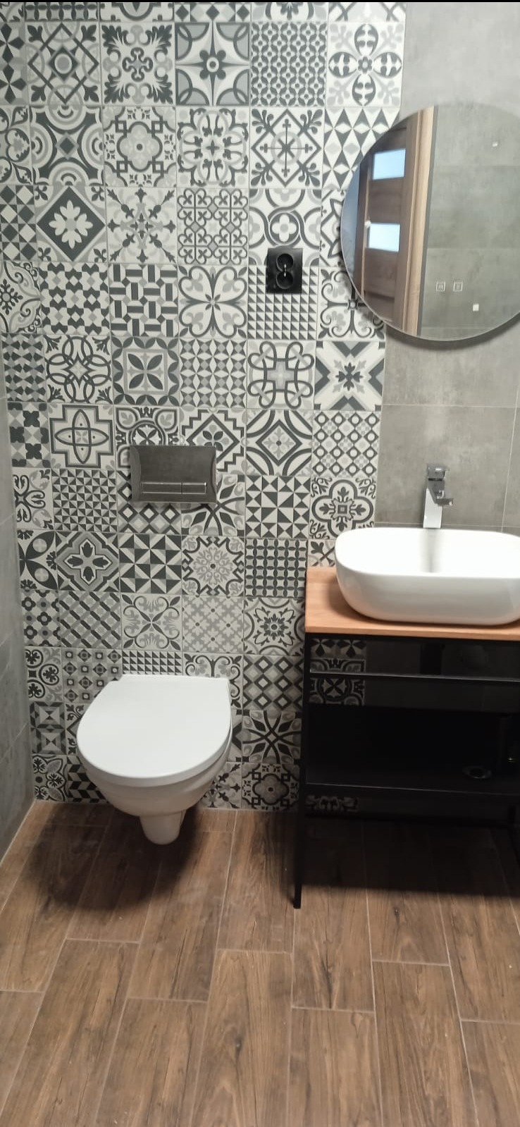 Ukończona łazienka z mozaiką na ścianie, okrągłym lustrem i drewnopodobną podłogą. Biała umywalka nablatowa na czarnej szafce. Nowoczesny design.