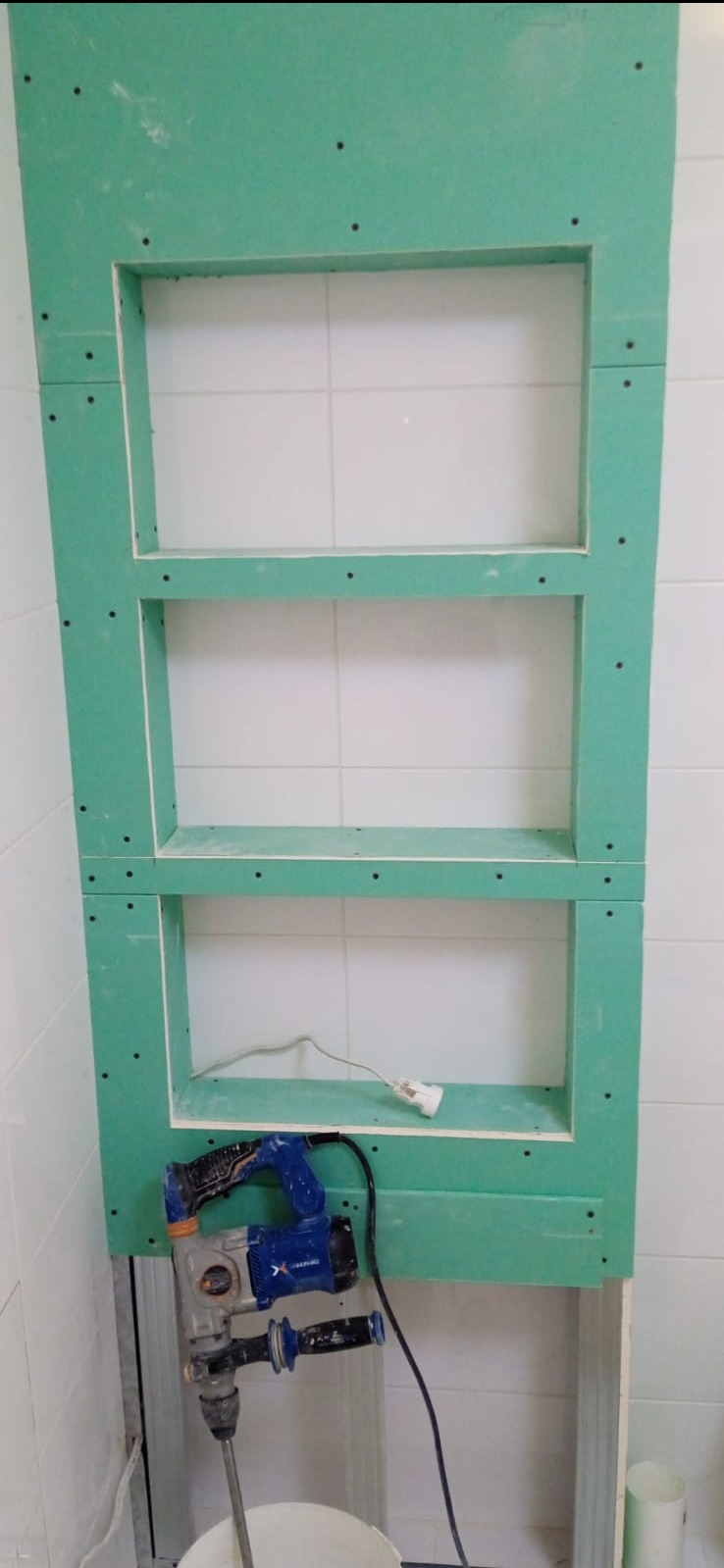 Nietypowa zabudowa z zielonej płyty GK z trzema wnękami na ścianie z białych płytek. Widoczny sprzęt budowlany i fragment podłogi w trakcie prac.