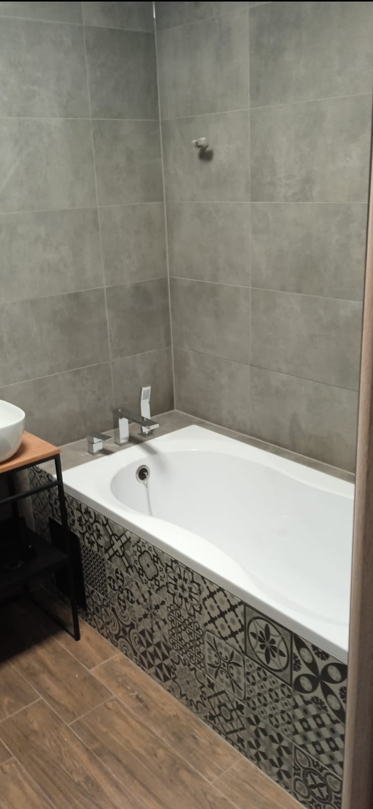 Wykończona łazienka z wanną obudowaną dekoracyjnymi płytkami w stylu patchwork, szare płytki na ścianach i drewnopodobna podłoga. Nowoczesna armatura.