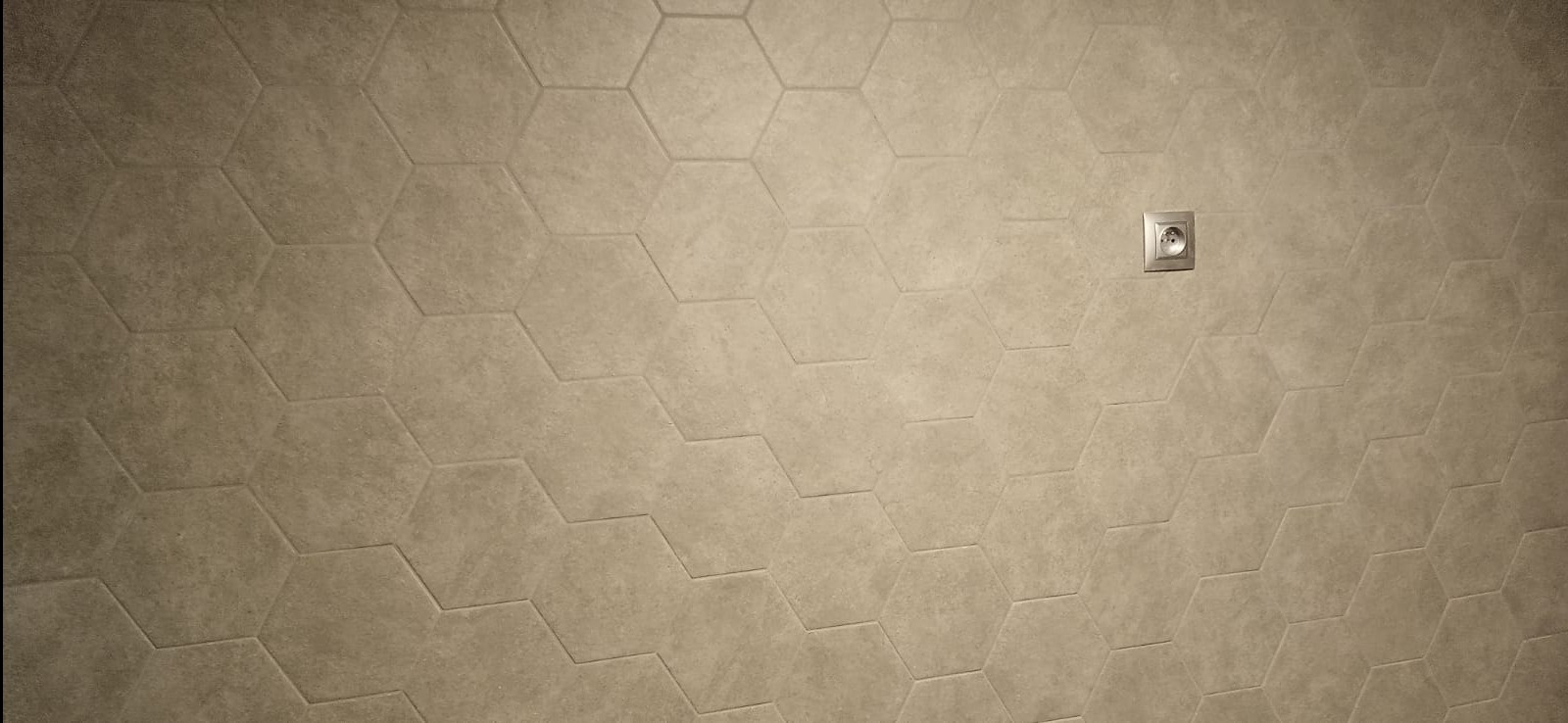 Beżowa ściana wyłożona heksagonalnymi płytkami ceramicznymi z widocznym gniazdkiem elektrycznym. Minimalistyczny design, neutralna kolorystyka.