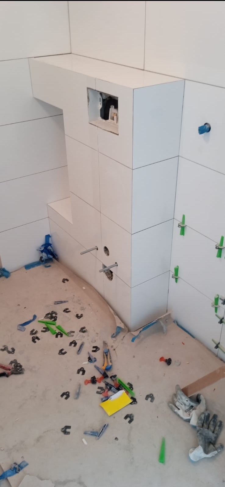 Białe płytki w łazience, częściowo ułożone, widoczne narzędzia glazurnicze i elementy montażowe. Otwór na instalację sanitarną.