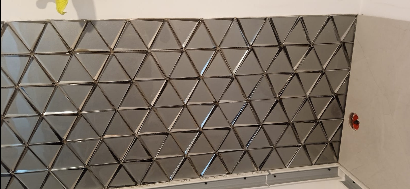 Ściana wyłożona błyszczącymi, trójkątnymi płytkami w odcieniach szarości, tworzącymi geometryczny wzór. Widoczny fragment ściany bez płytek i puszka elektryczna w Brwinowie.