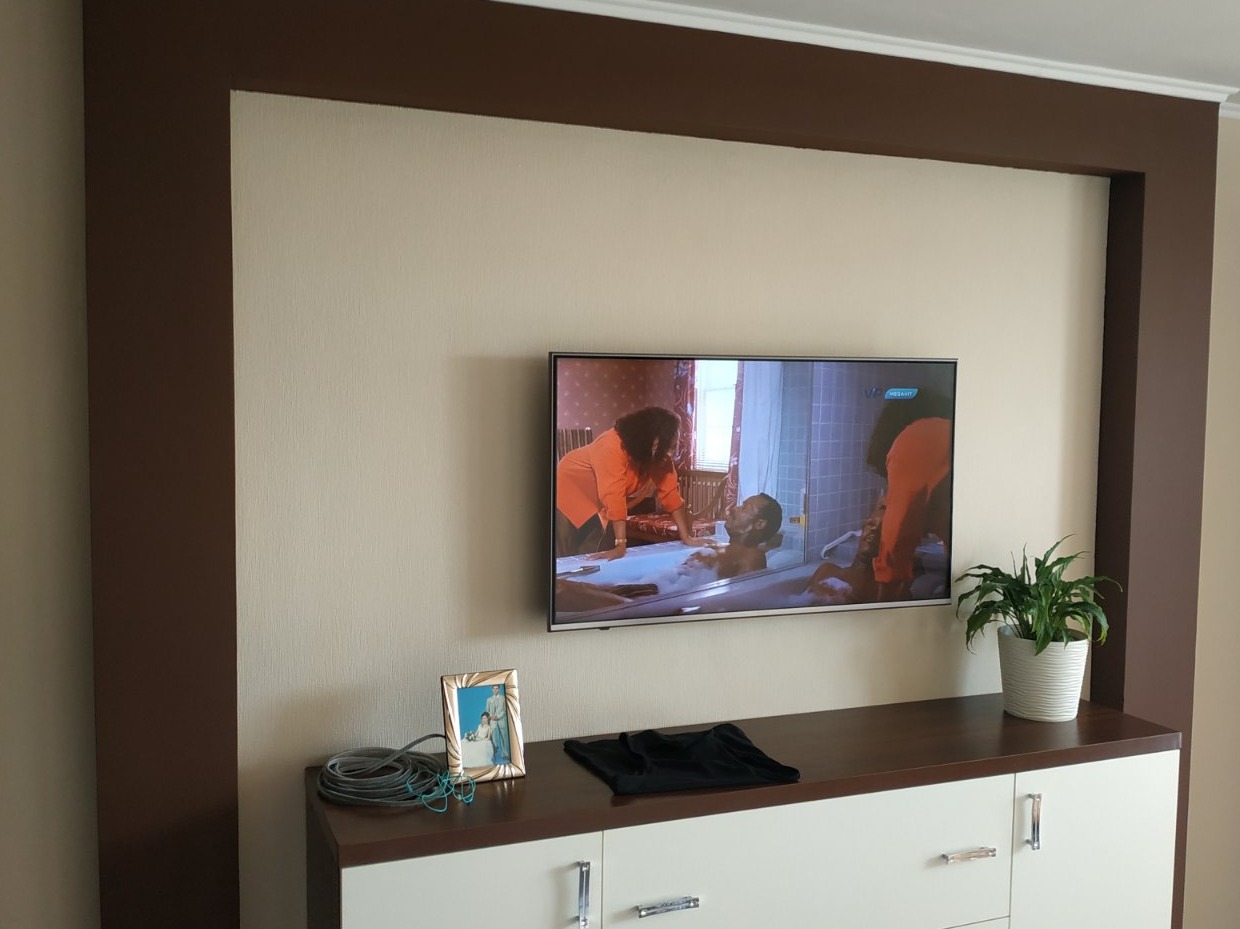 Ściana z beżową tapetą i brązową ramą, na niej telewizor. Poniżej biała komoda z brązowym blatem, na której stoi ramka ze zdjęciem i roślina doniczkowa.