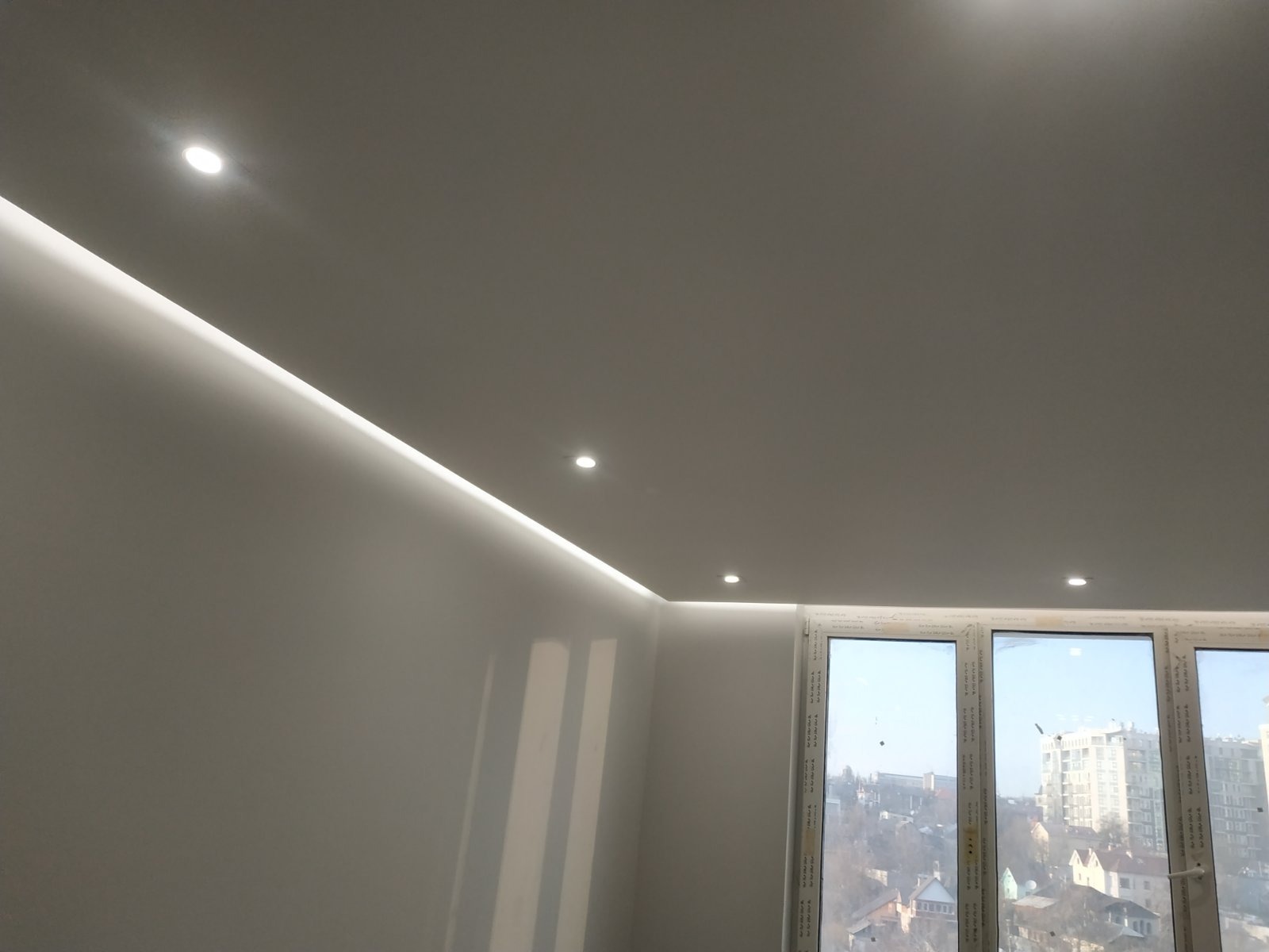 Biały sufit z oświetleniem LED wzdłuż krawędzi, widok na okno z widokiem na miasto w tle. Minimalistyczny design wnętrza, nowoczesne wykończenie.