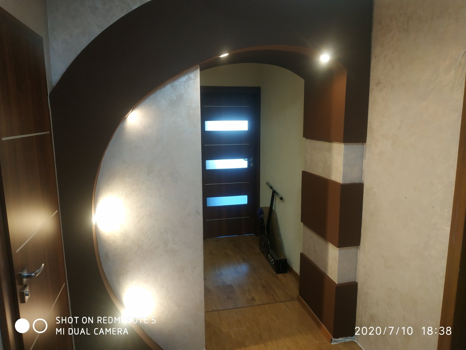 Nowoczesne wykończenie wnętrza: łuk z oświetleniem punktowym, dekoracyjna struktura ścian, drewniane drzwi i podłoga. Widoczny skuter w tle.