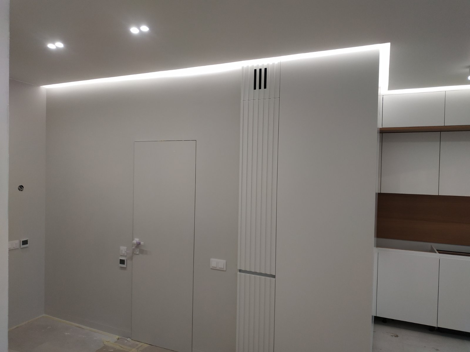 Minimalistyczne wnętrze po remoncie: białe ściany z ukrytymi drzwiami, dekoracyjny panel ścienny z pionowymi żłobieniami i oświetlenie LED podsufitowe.